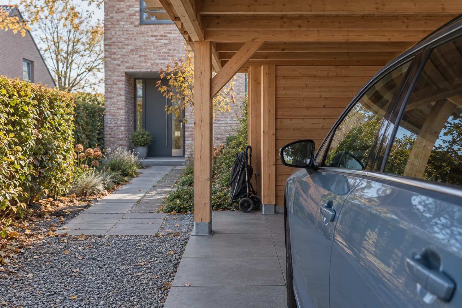 Voiture sous un carport en bois, poussette pliée à l’arrière et chemin dégagé vers la maison