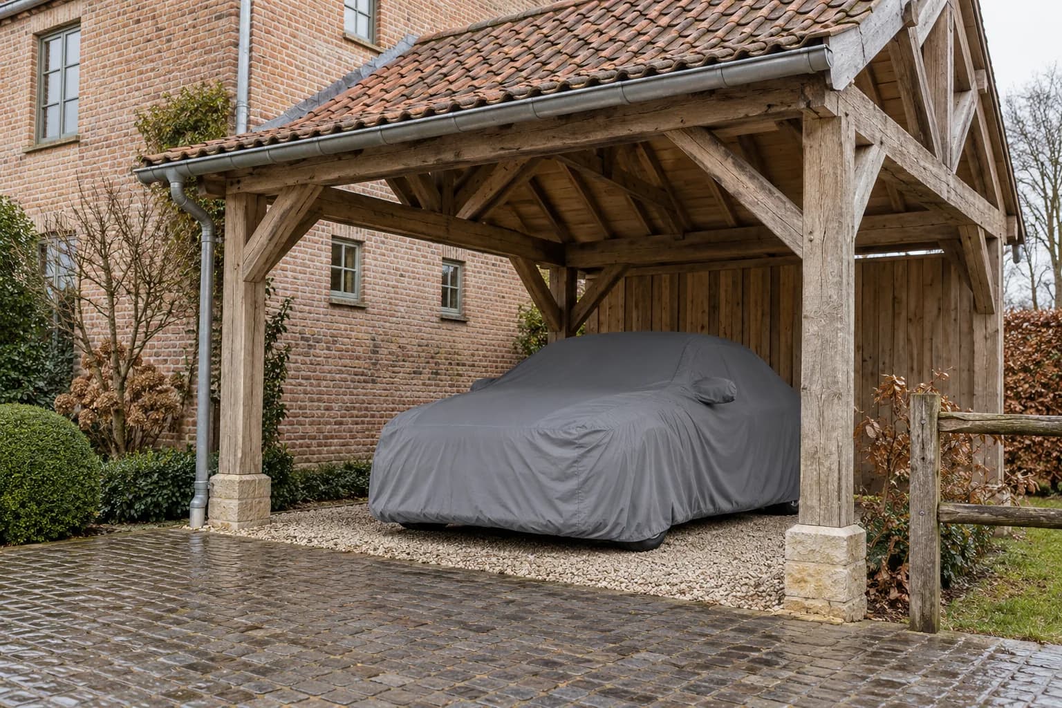 Voiture sous une housse grise dans un carport en bois à toit de tuiles, près d'une maison en briques.