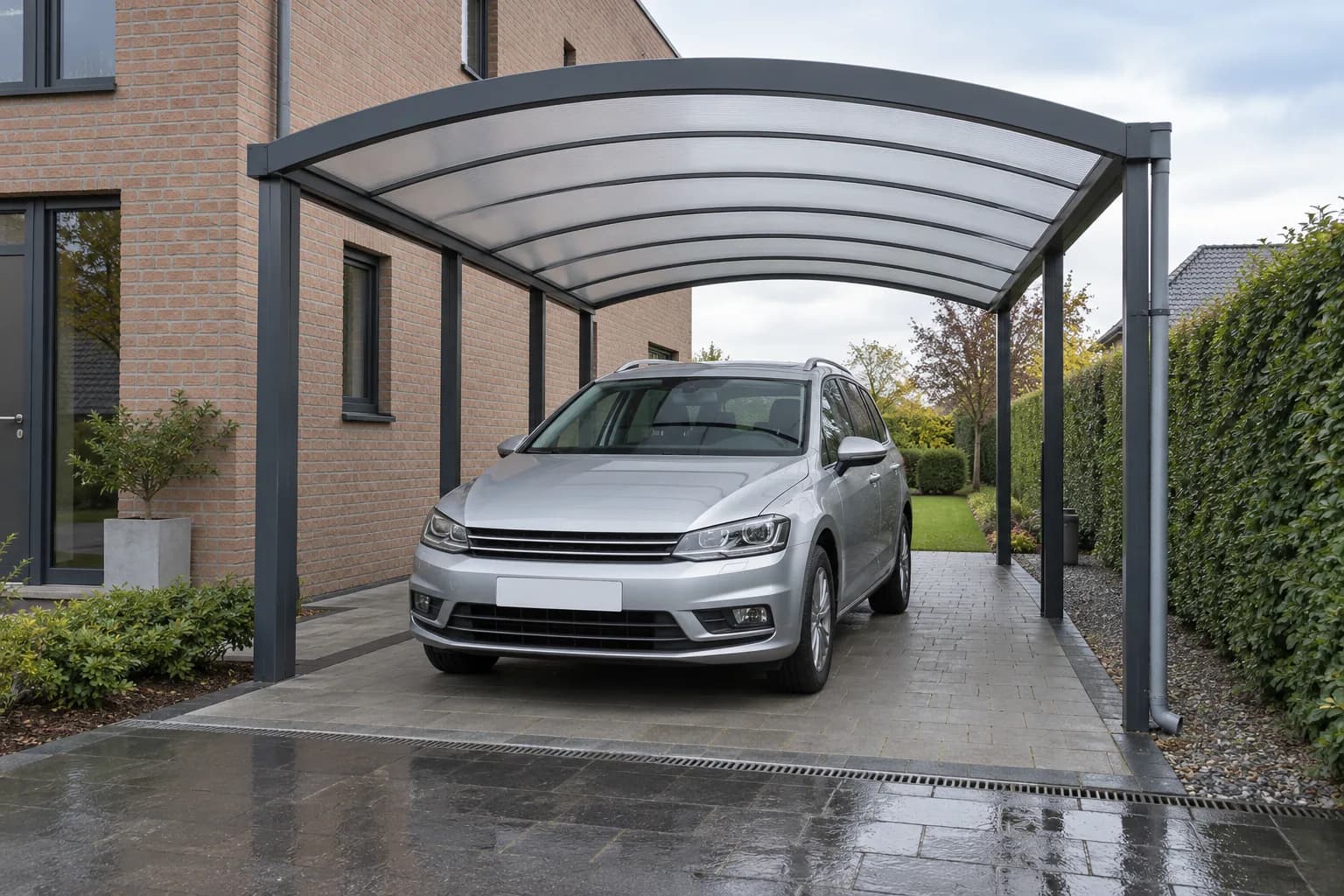 Voiture argentée garée sous un carport à toit arrondi, avec une allée humide devant l'abri.