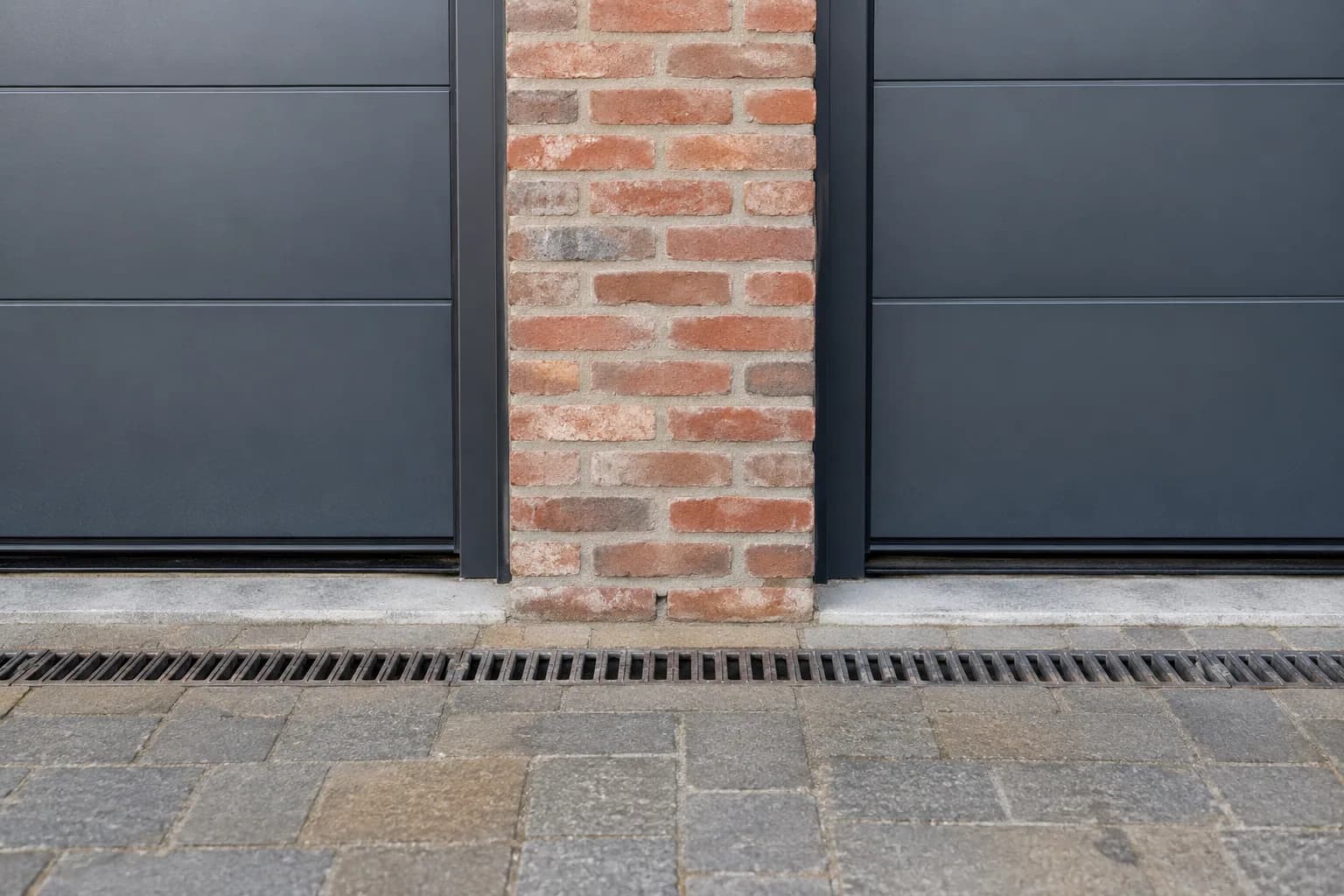 Pilier en briques entre deux portes de garage, avec joints de seuil et caniveau devant les ouvertures.