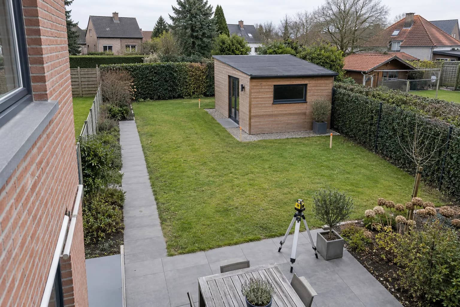 Vue sur une annexe indépendante dans un jardin clôturé, avec repères au sol et appareil de mesure sur trépied.
