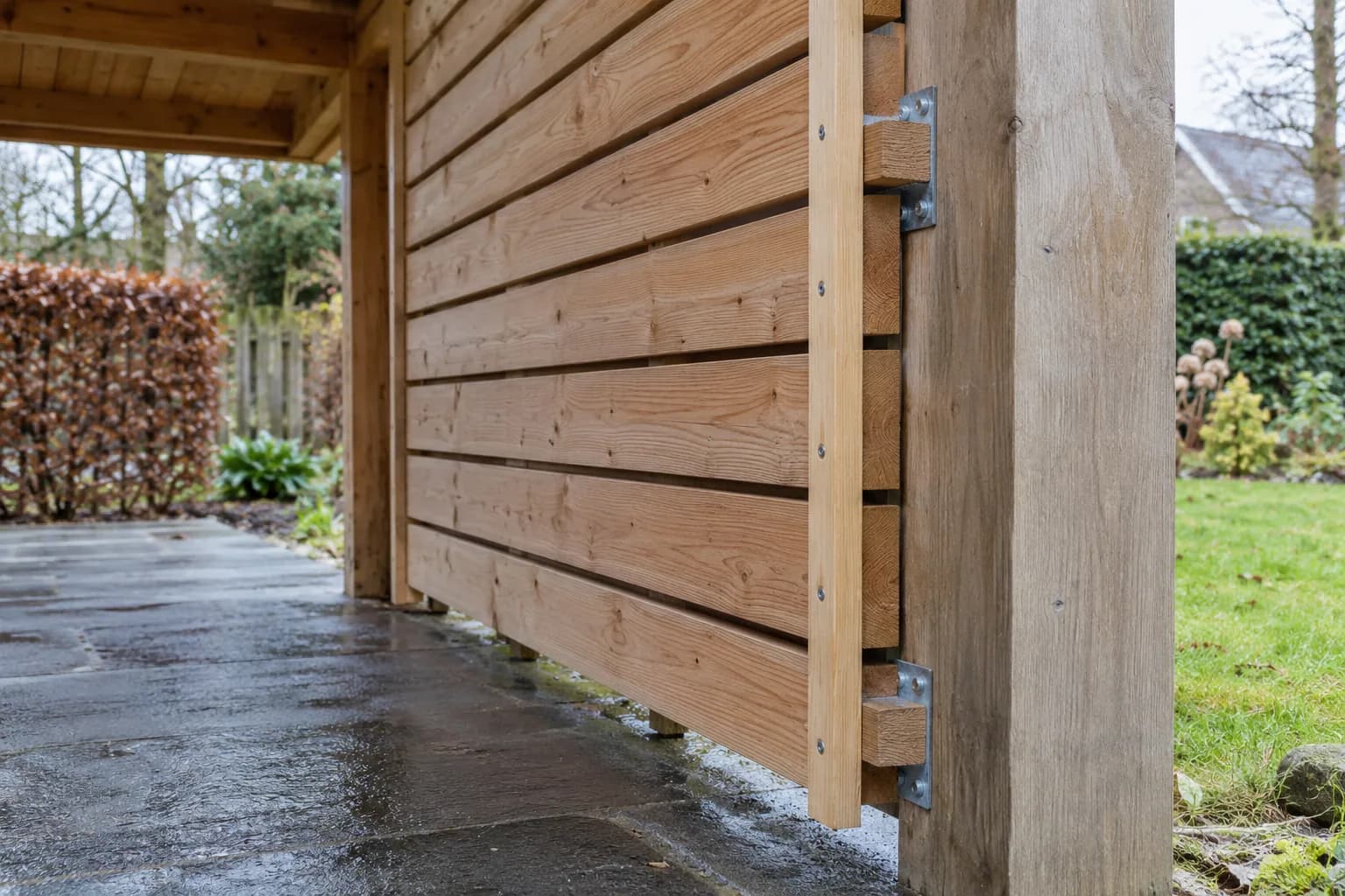 Fixations métalliques du bardage latéral sur un poteau de carport, avec un dégagement au-dessus du dallage.
