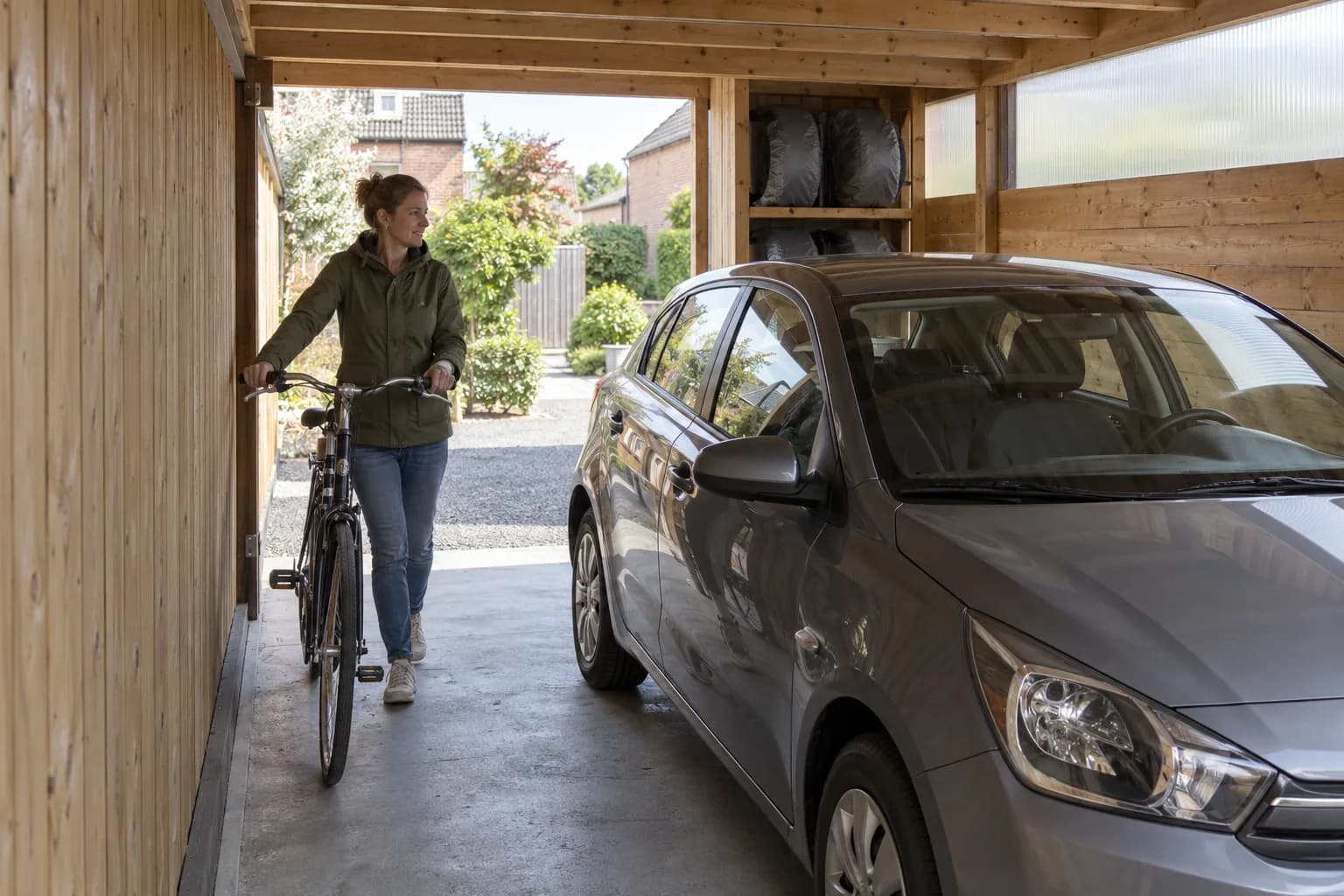 Une personne pousse son vélo dans le passage laissé entre une voiture et la paroi en bois du carport.
