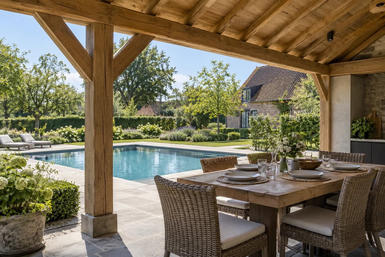 Table dressée sous la charpente en chêne d’un pool house, face à la piscine et au jardin.