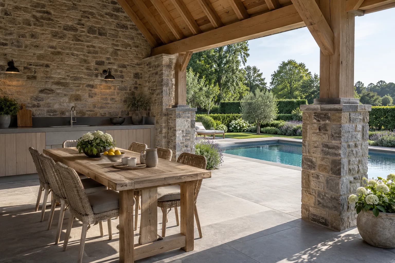 Cuisine d’été et table sous une toiture portée par des piliers maçonnés, dans un pool house en pierre.