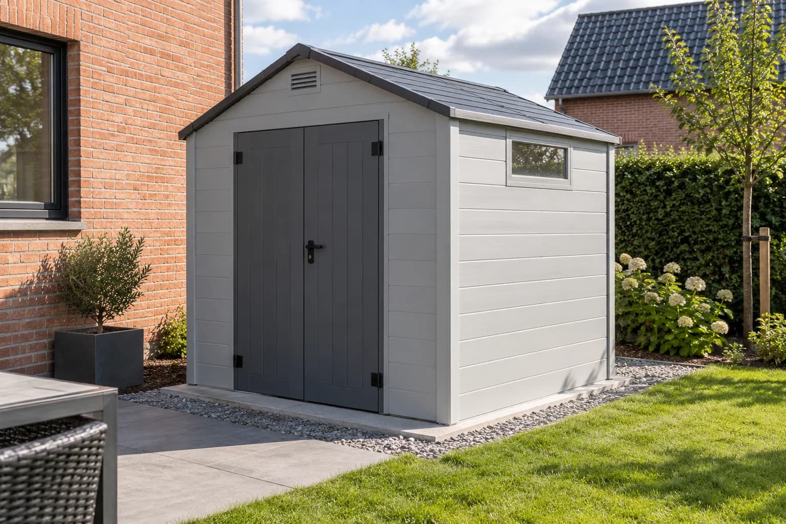 Remise en panneaux plastiques gris clair avec doubles portes sombres et grille d’aération, sur une dalle de jardin.