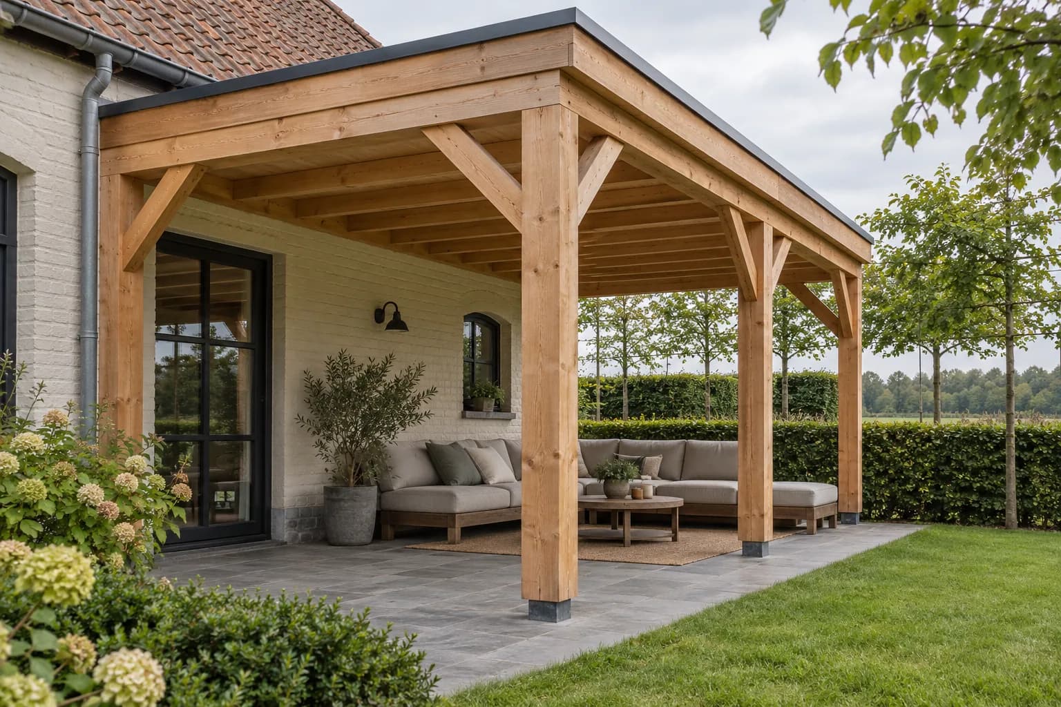 Terrasse couverte par une charpente en bois avec jambes de force, devant une maison en briques peintes.