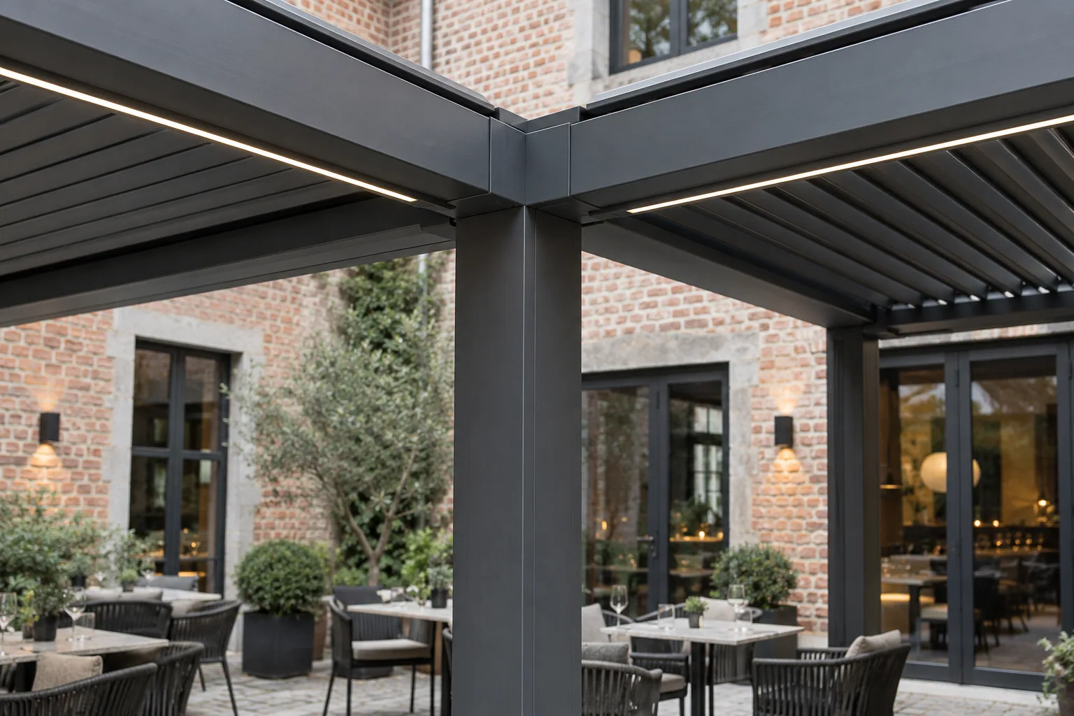 Jonction de poutres et d’un poteau sous une toiture de terrasse, avec éclairage intégré et tables en arrière-plan.
