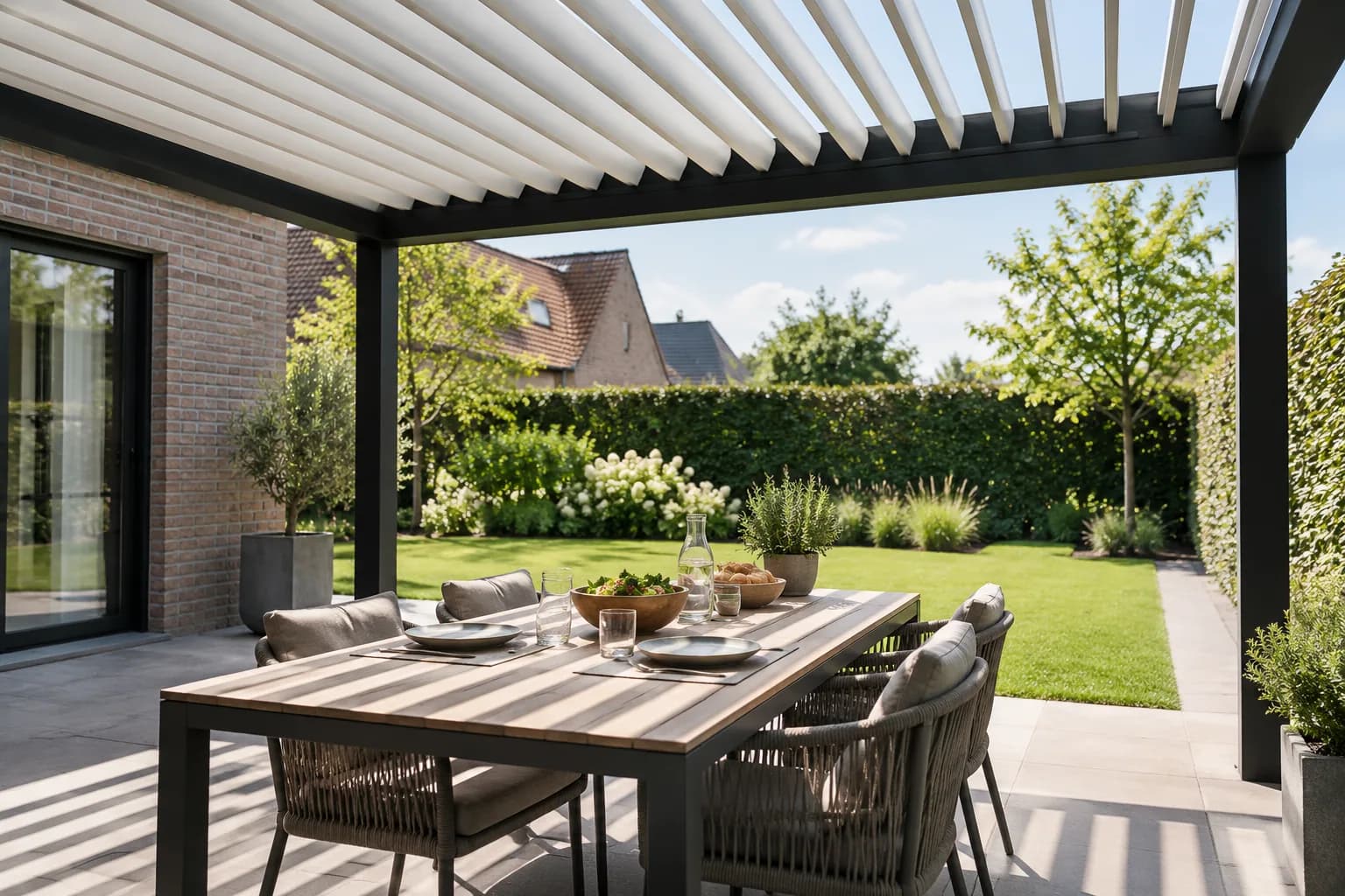 Table dressée sous les lames orientables d’une pergola, avec des bandes d’ombre sur la terrasse.