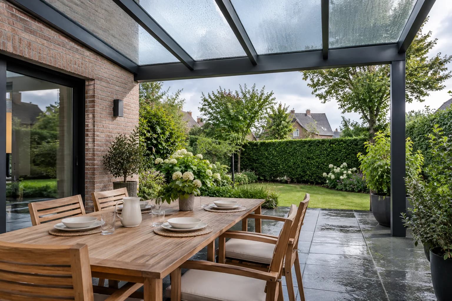Table dressée sous une toiture vitrée, avec terrasse mouillée et jardin visibles au-delà de l’abri.
