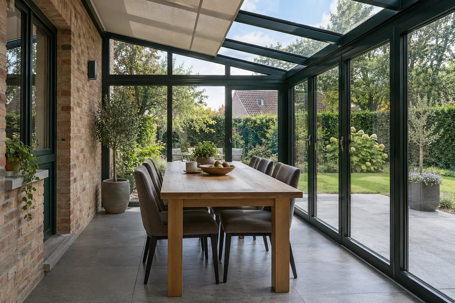 Salle à manger dans une véranda vitrée aux châssis vert foncé, avec un store sous la toiture.