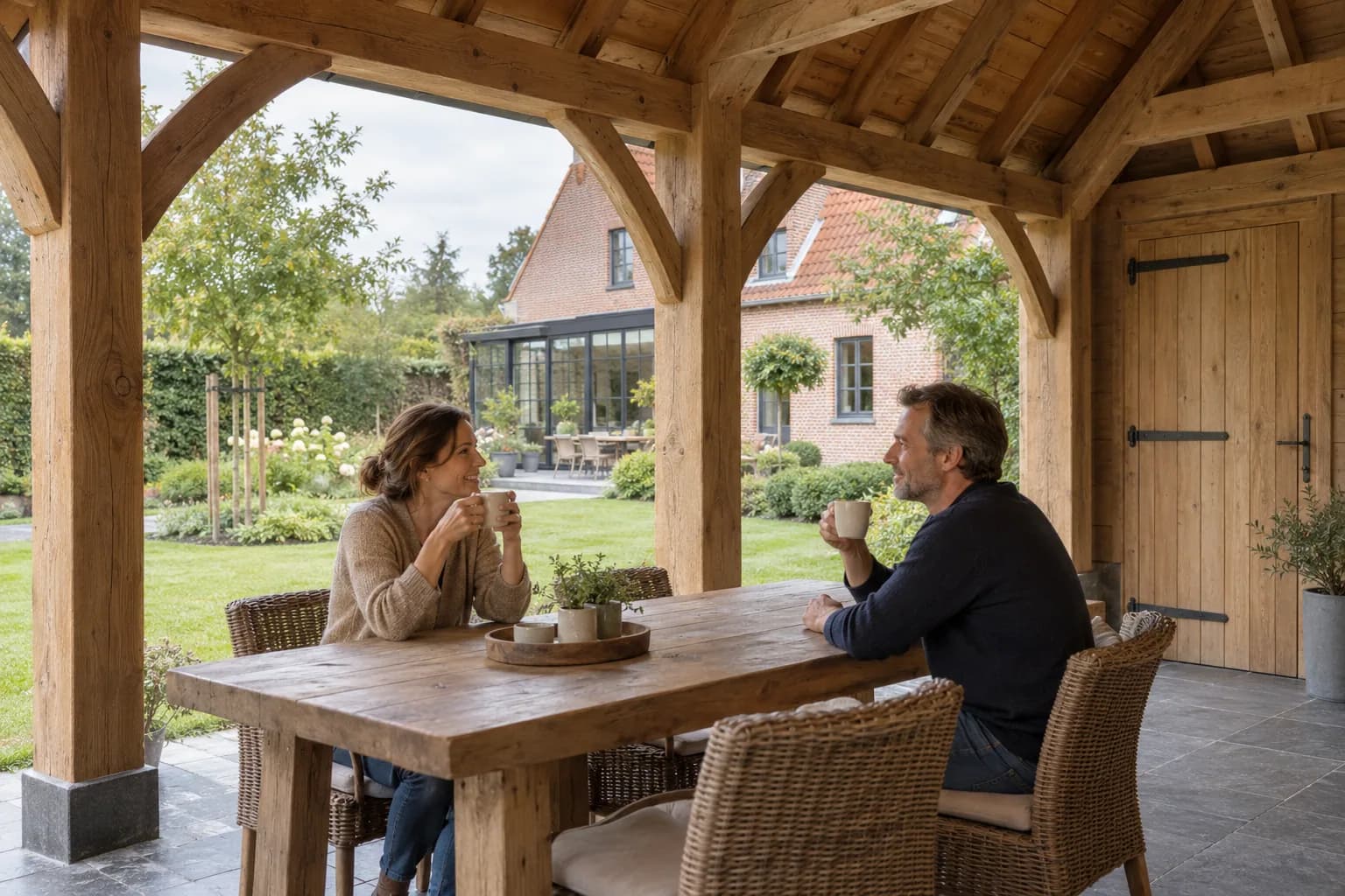 Twee bewoners zitten met kopjes aan een houten tafel onder het zichtbare eiken gebinte van een bijgebouw.