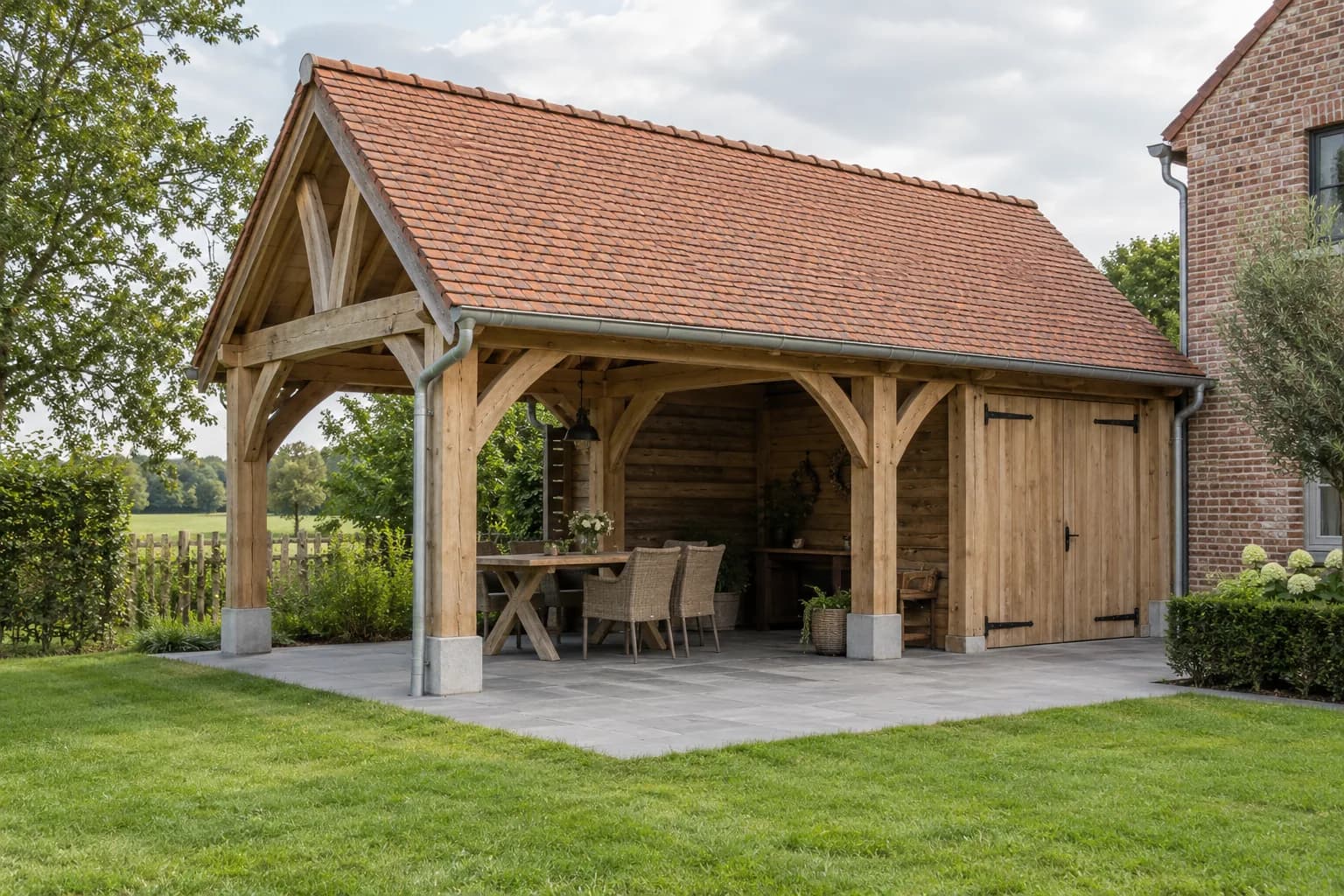 Eiken bijgebouw met pannendak, gebogen schoren, overdekte eethoek en afgesloten berging naast een bakstenen huis.