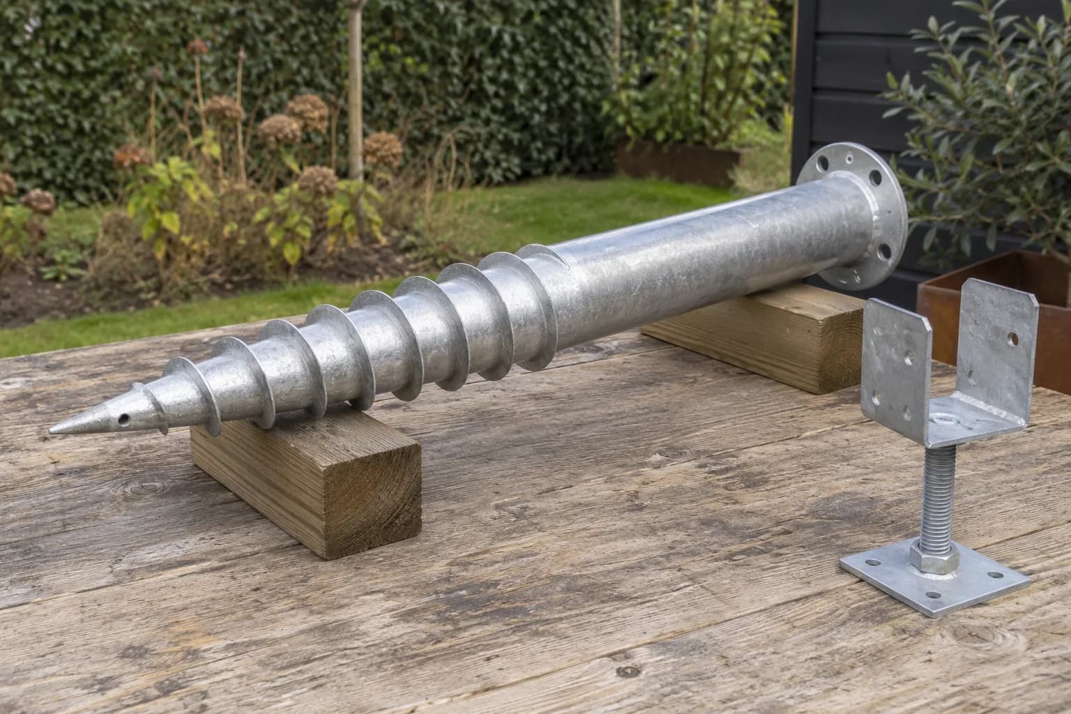 Verzinkte grondschroef met spiraal en flens, naast een verstelbare balkdrager op een houten werktafel
