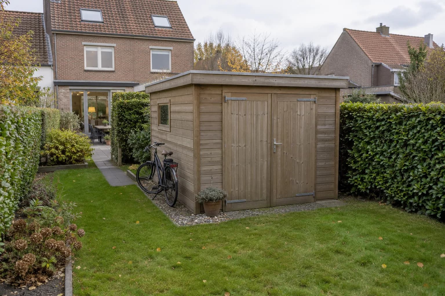 Compact houten tuinhuis met dubbele deuren en een fiets naast het pad in een smalle achtertuin