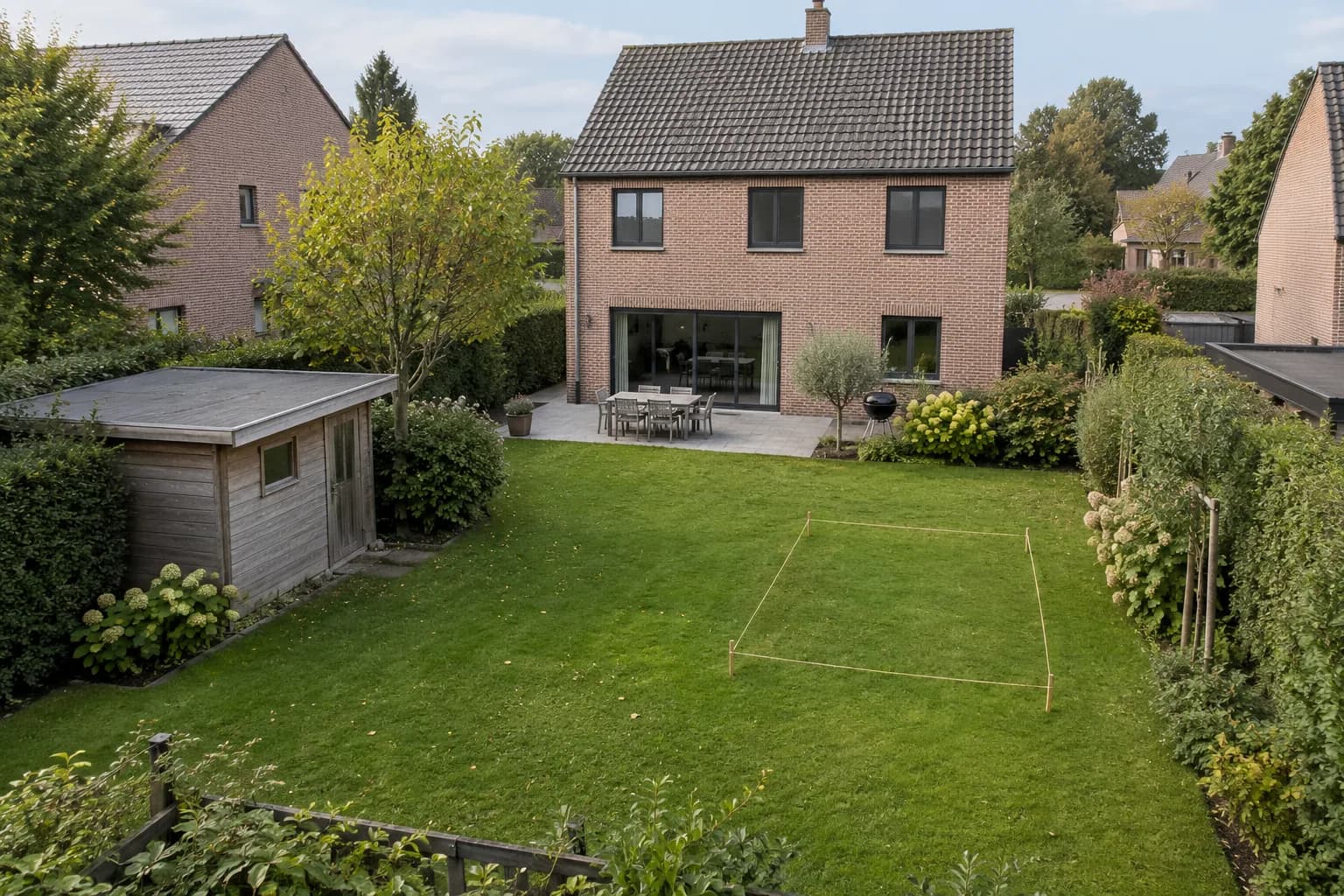 Bestaand tuinhuis en met koord uitgezette bouwplek in een omhaagde tuin achter een bakstenen woning