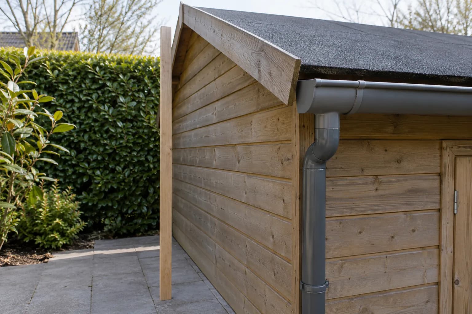 Dakrand van een houten tuinhuis met roofing, dakgoot en regenpijp, naast een rechtopstaande houten lat