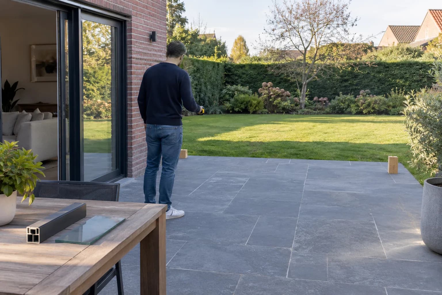 Bewoner bekijkt de terrasruimte met een meettoestel; op tafel liggen een aluminiumprofiel en een glasstaal