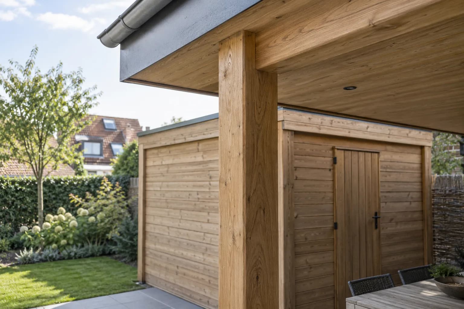 Detail van een houten staander en dakbalk naast de berging van een tuinhuis met overkapping.